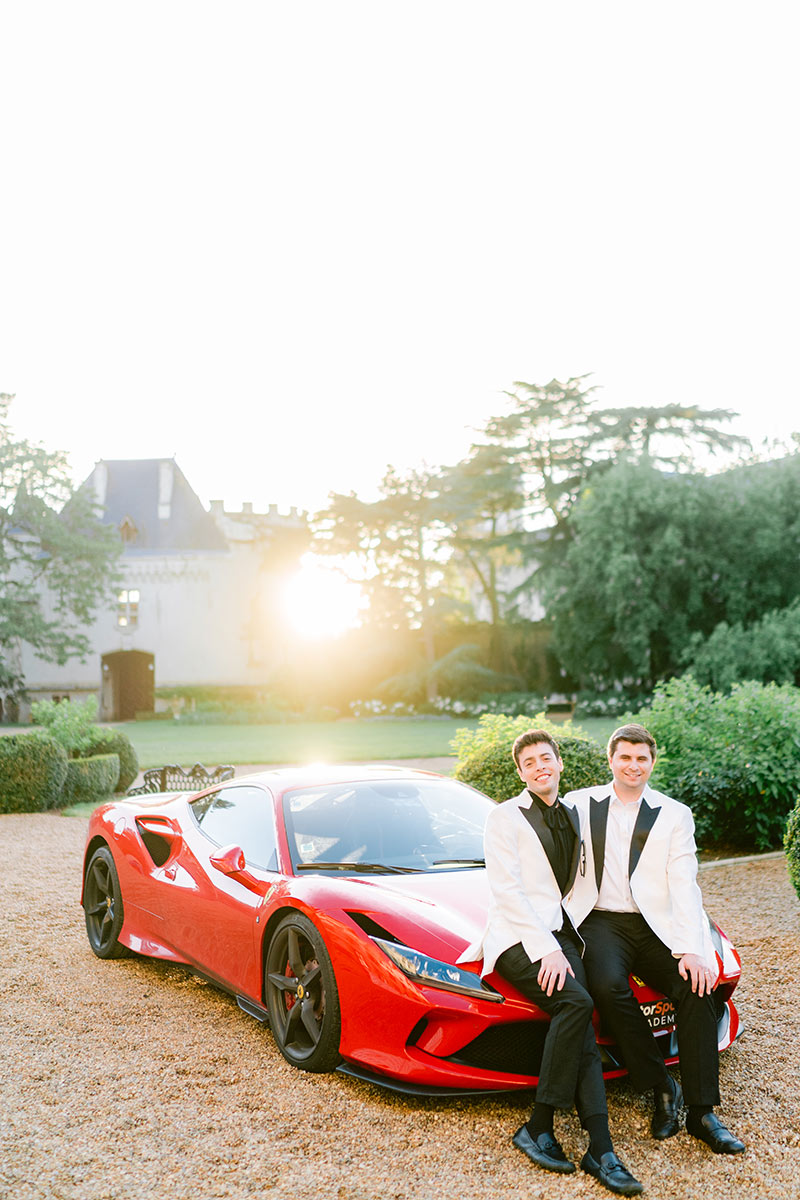 Two grooms sit on a red sports car after their luxury wedding celebration at Chateau Challain in The Loire Valley, France by French Wedding videographer Emma Wilson from Story Of Your Day