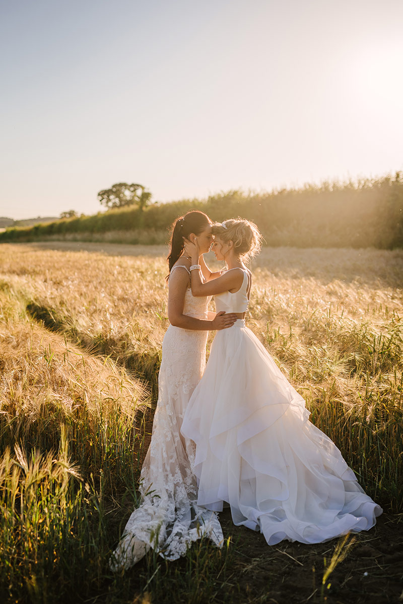 Two women in wedding dresses stand closely together in a sunlit field, sharing an intimate moment—perfect for capturing on a stunning wedding video by a UK wedding videographer.
