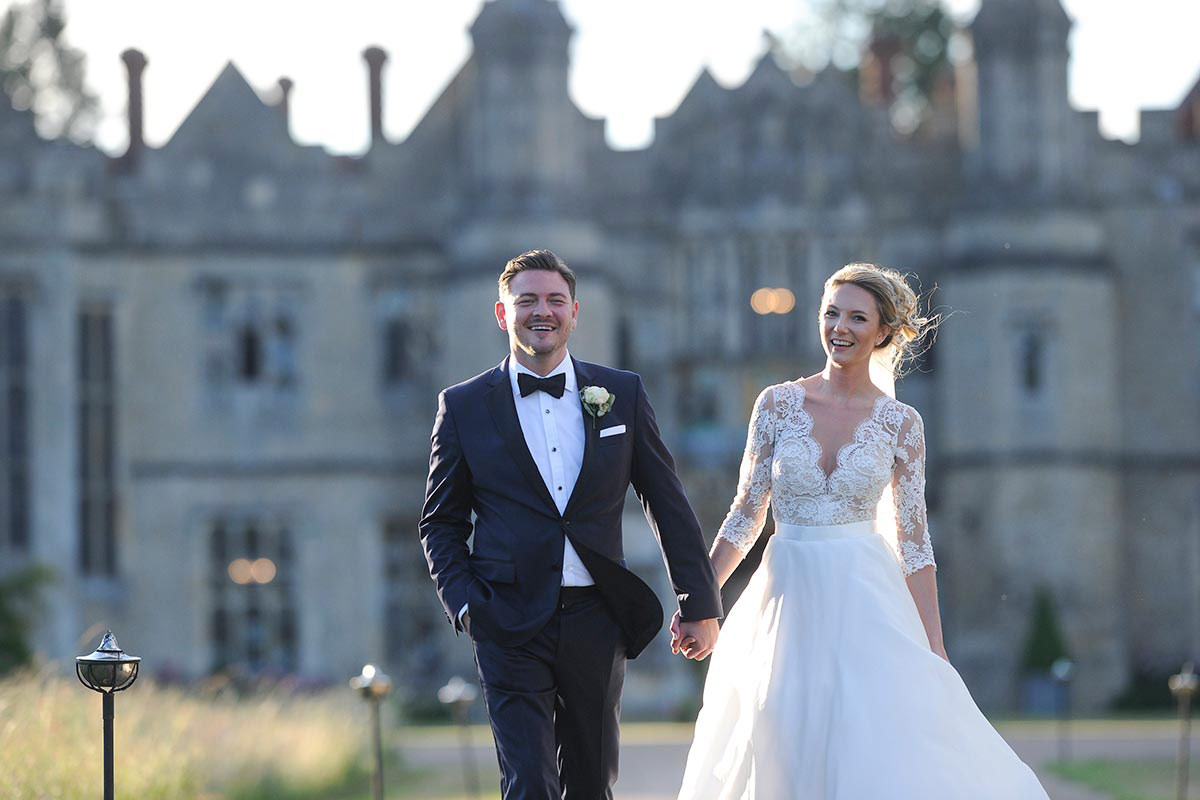 A bride and groom in formal attire walk hand-in-hand outdoors, smiling, with Hengrave Hall in the background—captured beautifully by a UK wedding videographer Emma Wilson