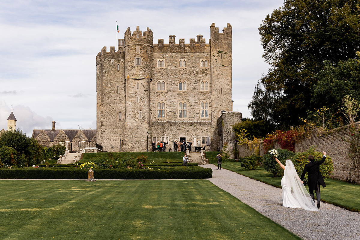 A bride and groom walk on a path toward a large stone castle with manicured lawns and gardens, as guests gather near the entrance—an enchanting scene perfect for UK Wedding Videography.