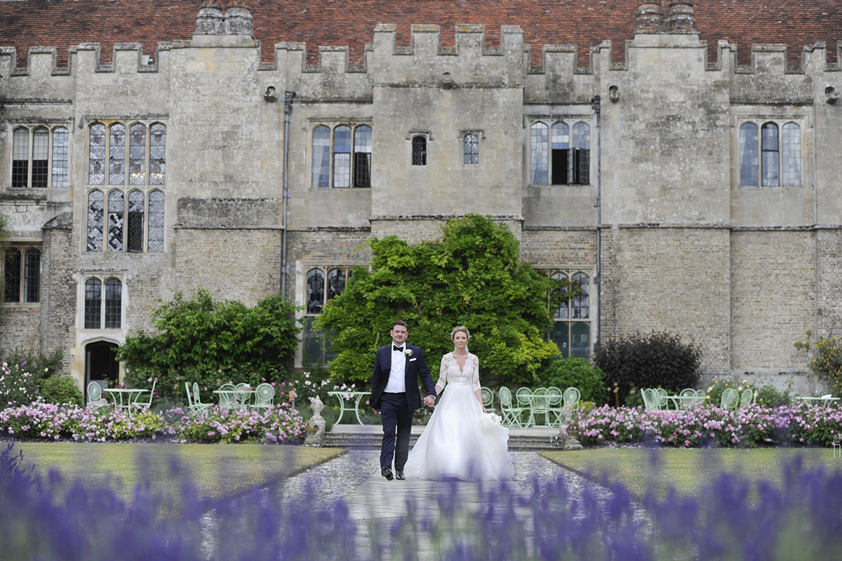 A bride and groom walk hand-in-hand down a garden path in front of a historic stone building, surrounded by flowers and greenery