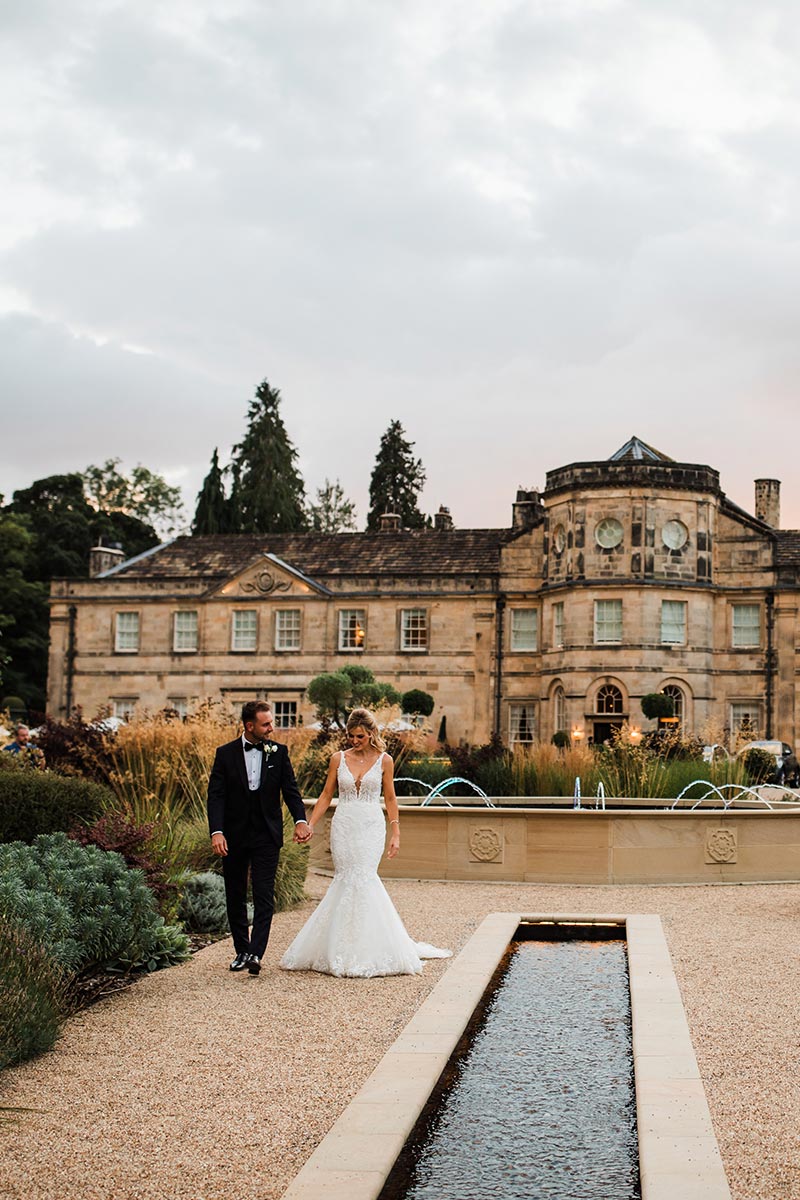 A bride and groom walk hand in hand along a gravel path in front of a grand historic building with fountains and landscaped gardens, perfectly captured by Story Of Your Day Wedding videographer