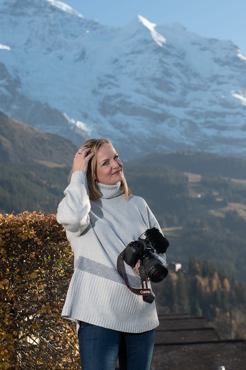Emma Wilson holds a Canon camera and touches her hair, standing outdoors with mountains and autumn foliage