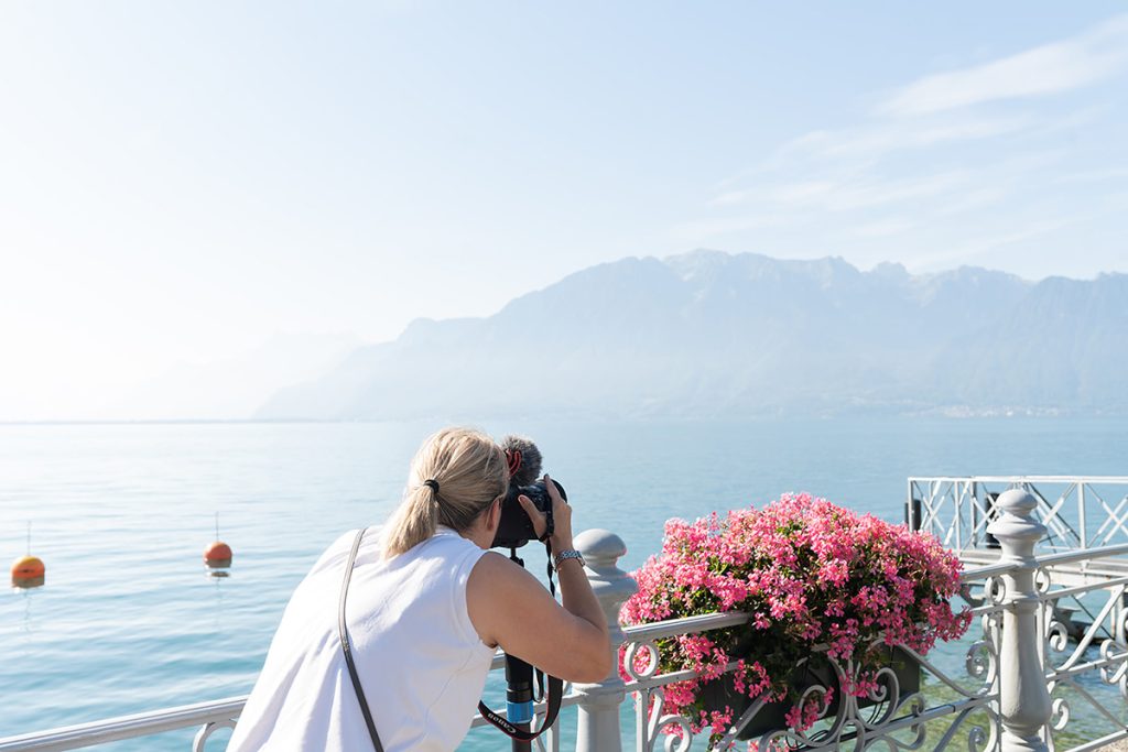 Emma Wilson from Story of Your Day leans over a railing decorated with pink flowers, filming a calm lake and distant mountains under a clear sky.