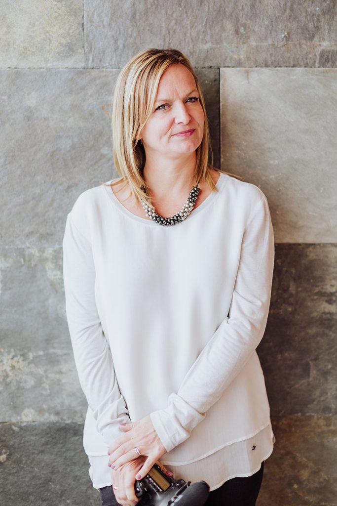 Emma WIlson, luxury wedding videographer stands in from of a stone tiled wall.