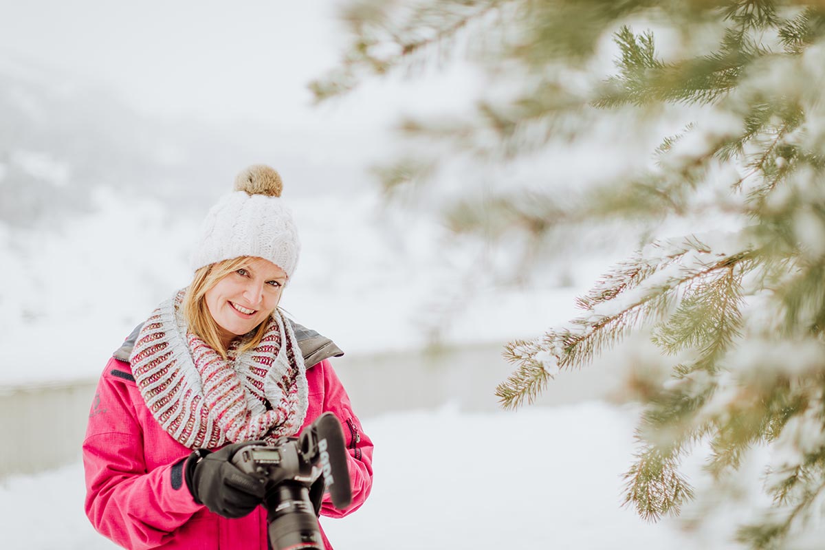 Emma Wilson, in a pink jacket and winter hat stands outdoors in a snowy Switzerland landscape, holding a camera and smiling near a pine tree.