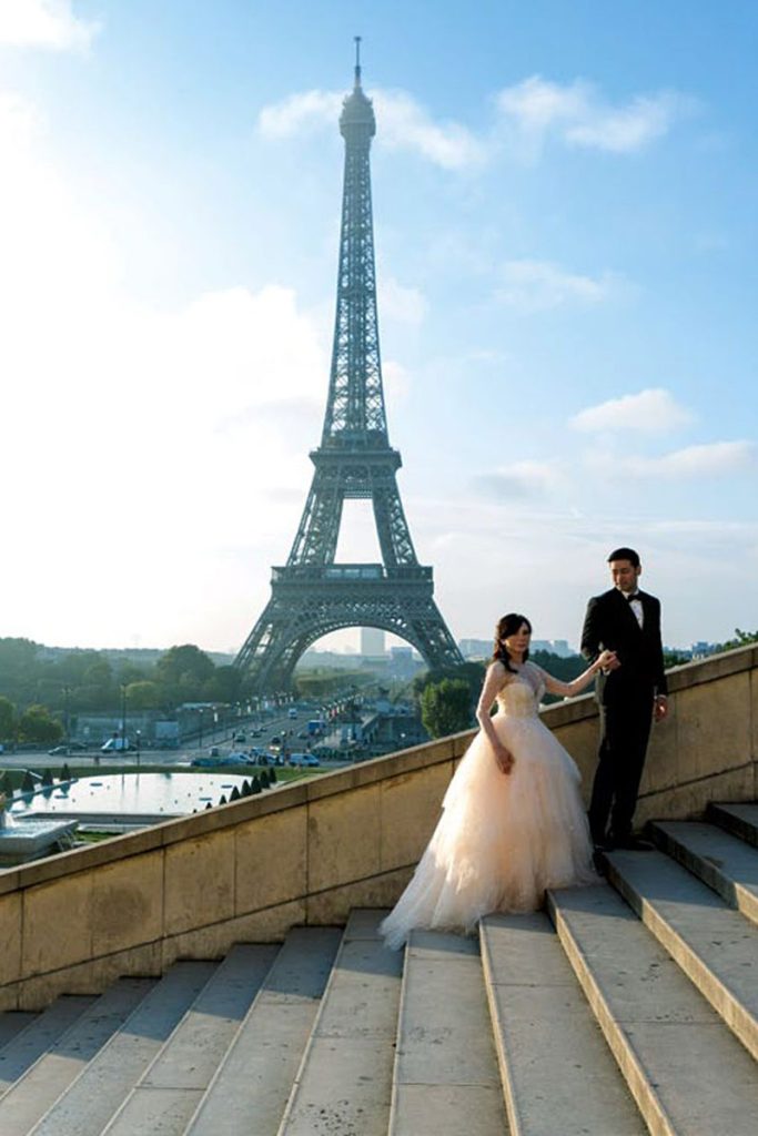A bride and groom hold hand walking up stairs with the Eiffel tower behind them after their wedding in Paris, France