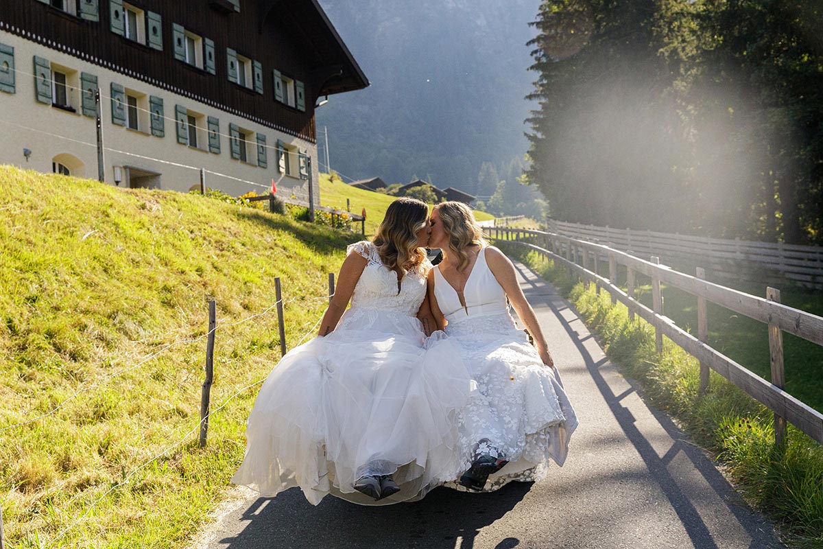 Two brides in white dresses sit on an old trailer, kissing, with a wooden house, grassy slope, and sunlight in the background—capturing the joy and beauty of LGBTQ+ weddings in an idyllic countryside setting.