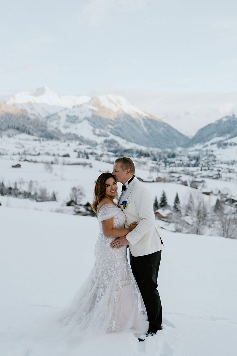 A bride and groom stand together in the snow in Switzerland, with mountains and a village in the background. The groom wears a white jacket and kisses the bride's forehead—perfect for capturing memorable wedding videography.