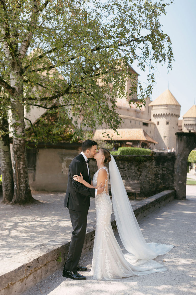 A bride and groom stand outside near a tree and stone wall, sharing a kiss in front of a historic building with towers—perfect for capturing timeless wedding videography in Switzerland.
