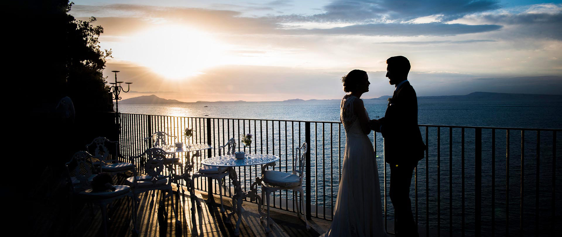 A couple stands on a balcony overlooking the sea at sunset in Italy, silhouetted against the sky with tables and chairs nearby