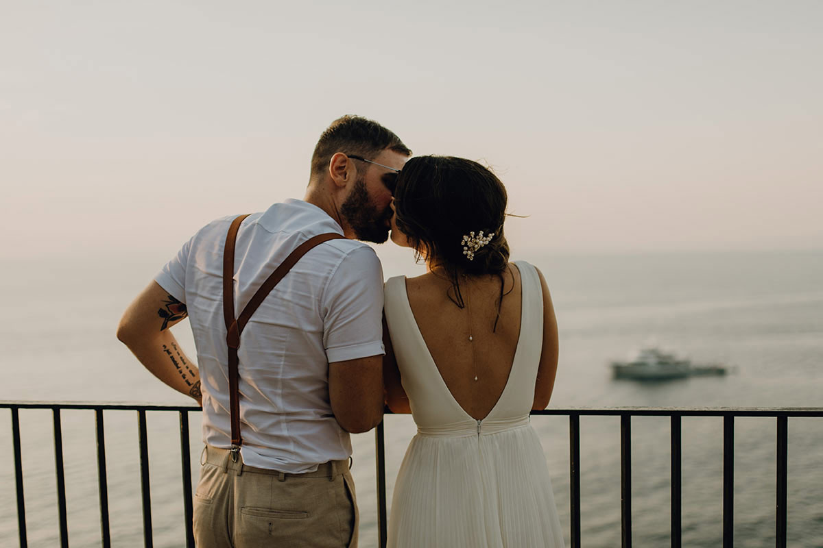 A couple stands on a balcony overlooking the sea in Italy, the man whispering to the woman in a white open-back dress. A boat glides below