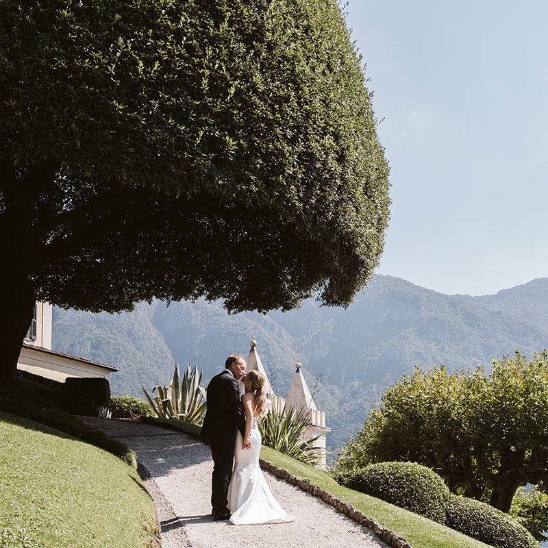 A bride and groom stand on a curved garden path, kissing beneath a large, sculpted tree with mountains in the background—an enchanting moment perfect for Wedding Videography Italy.