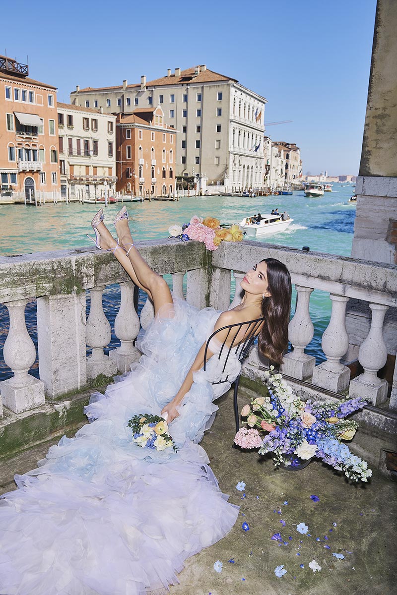 A woman in a ruffled gown reclines on a balcony overlooking an Italian canal, holding a bouquet of flowers with more scattered nearby. Boats and historic buildings set the scene