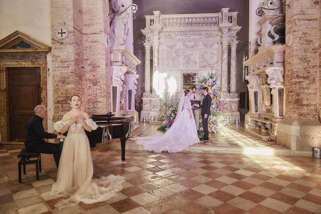 A bride and groom stand at the altar during a beautiful wedding ceremony in Italy, as a pianist and masked singer perform nearby