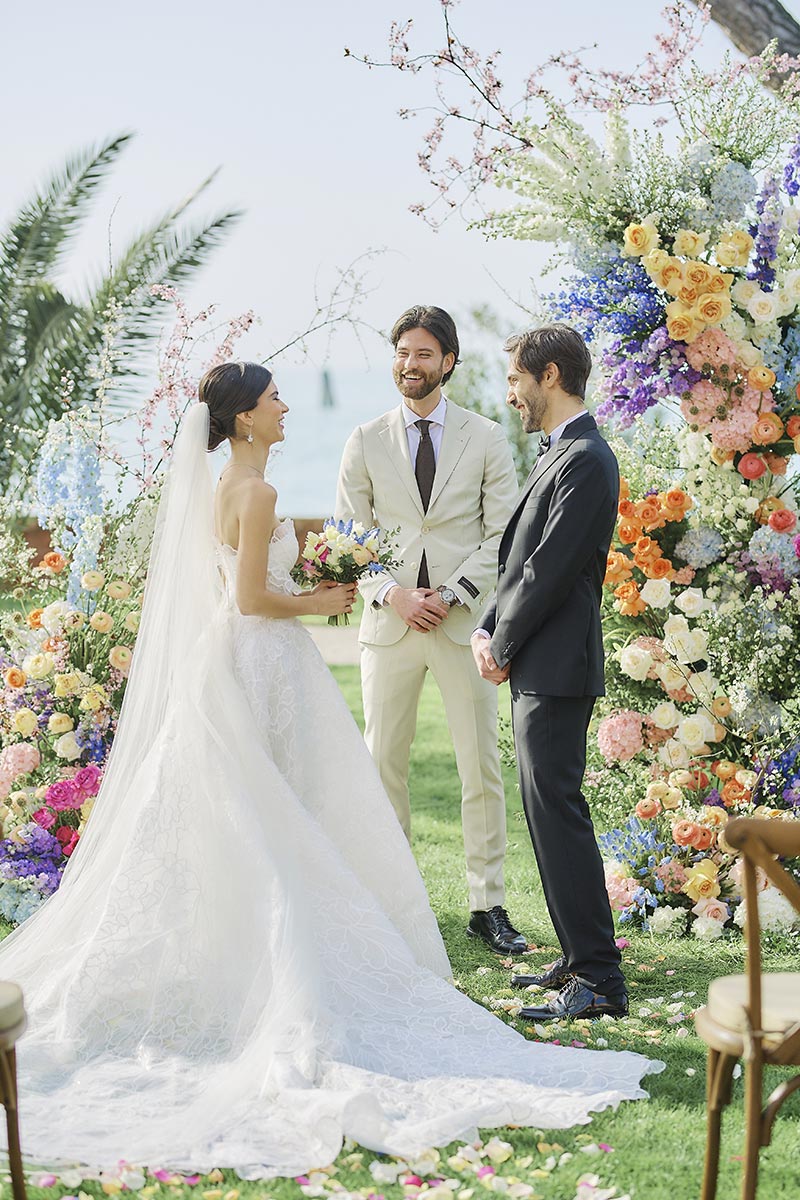 A bride and groom stand facing each other during an outdoor Italy wedding ceremony, with a smiling officiant and vibrant floral arrangements in the background—perfect moments to capture on wedding video.
