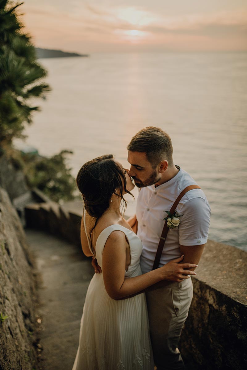A bride and groom embrace and kiss on a stone pathway beside the sea at sunset, perfectly capturing the romance of wedding videography in Italy.