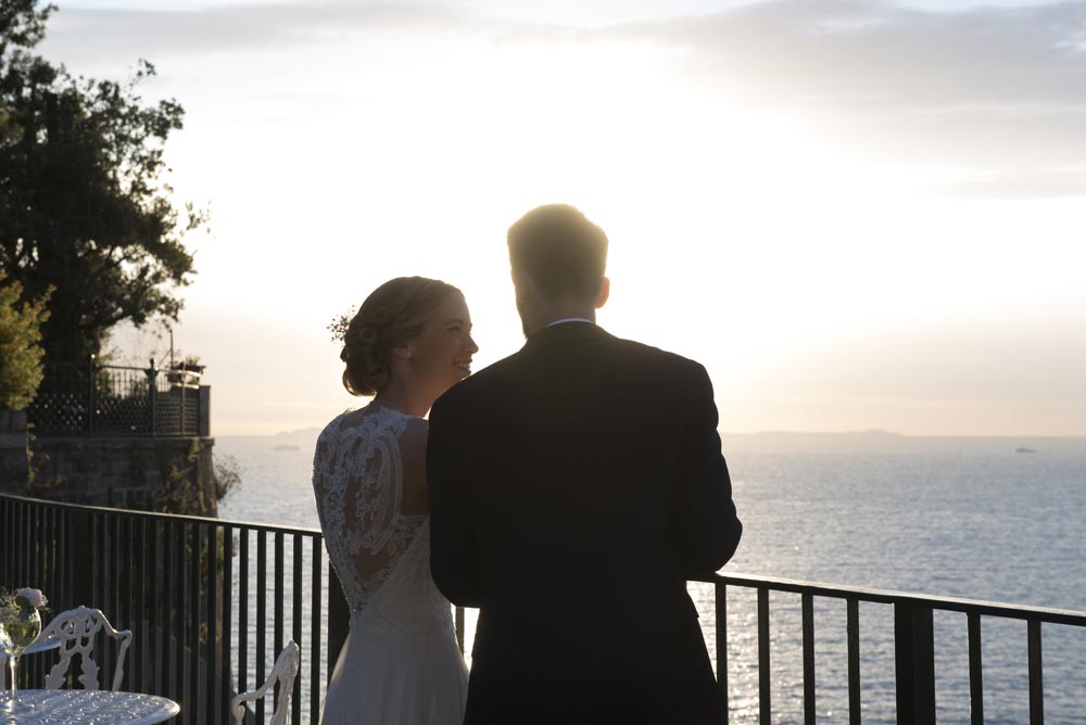 A bride and groom stand together on a balcony overlooking the ocean at sunset, their backs partially turned to the camera—a perfect moment for capturing exquisite wedding videography in Italy.