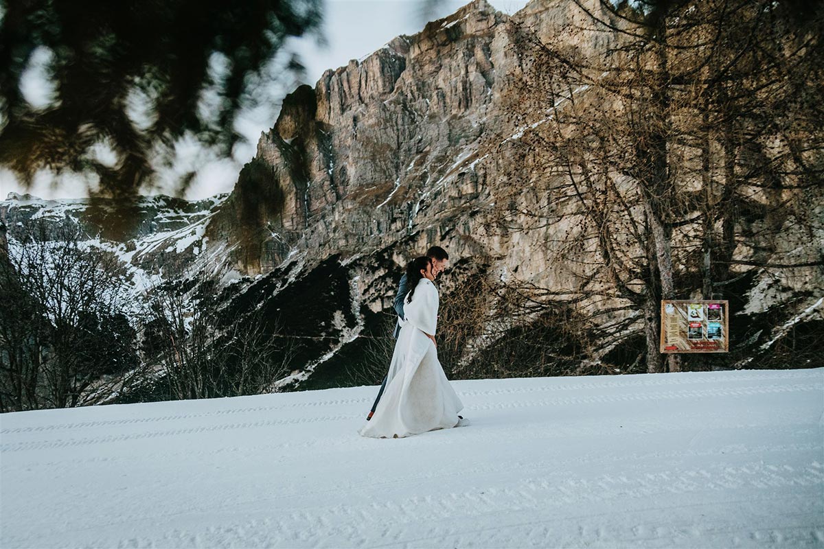 A bride in a white dress and her groom walks on snow in front of a rocky, mountainous landscape with sparse trees and a sign on the right