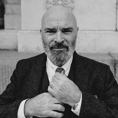 A bald man with a beard adjusts his tie while wearing a suit, standing outdoors in front of a stone building.