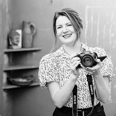 A woman smiling and holding a Canon camera; shelves with pottery and a bust sculpture are visible in the background.