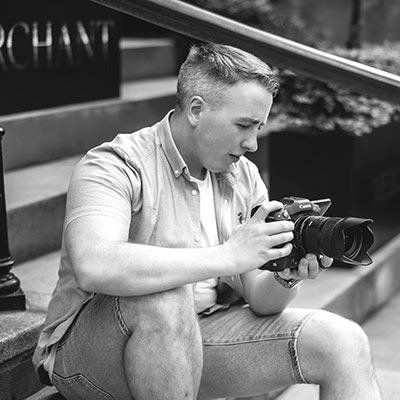 A person sitting on outdoor steps examines a DSLR camera, wearing a short-sleeved shirt and shorts. The scene is in black and white.