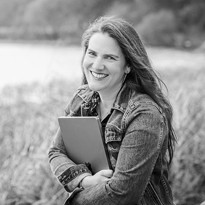 A woman with long hair wearing a denim jacket smiles at the camera while holding a closed laptop outdoors near tall grass.
