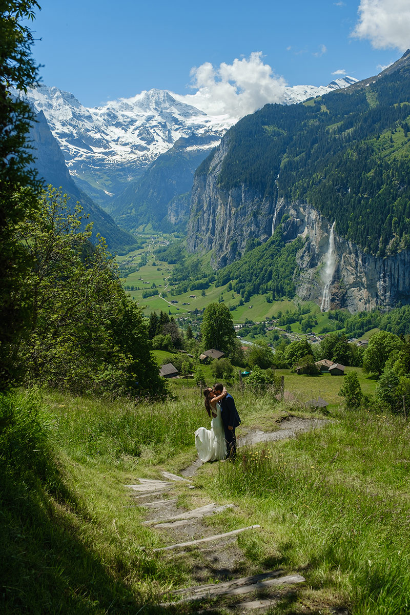 Un couple de mariés se tient sur un chemin herbeux surplombant la vallée de Lauterbrunnen, avec des montagnes enneigées et une cascade en arrière-plan : une scène parfaite pour un vidéaste spécialisé dans les fugues amoureuses sous un ciel bleu.