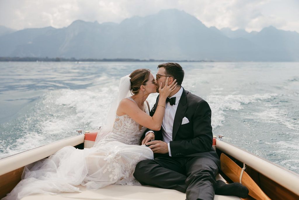 A bride and groom sit on a boat in Switzerland, dressed in wedding attire, kissing with mountains and water in the background—an unforgettable moment perfect for wedding videography.