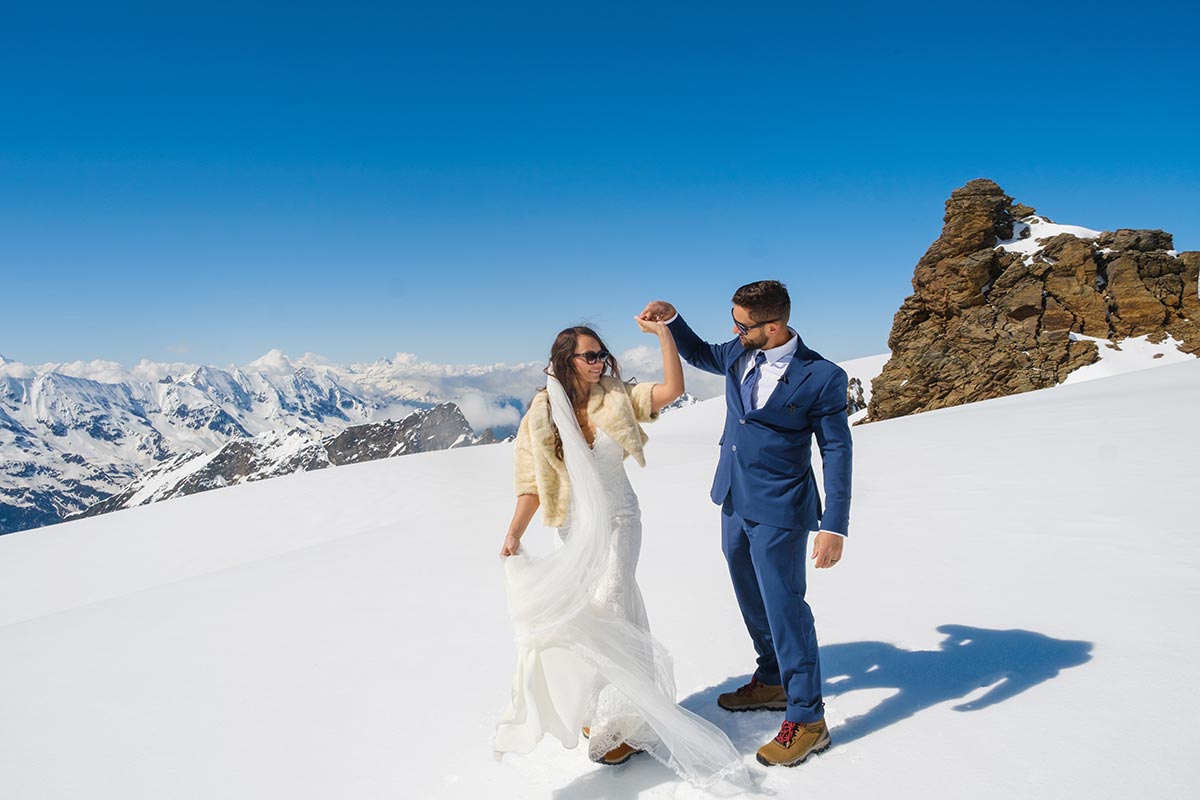 A bride and groom dance on a snowy mountain slope in Switzerland under a clear blue sky, with rocky peaks and snow-covered mountains in the background—a magical scene perfect for wedding videography.