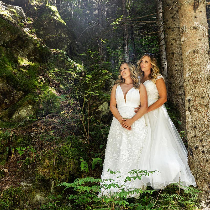 Deux mariées en robe blanche se tiennent côte à côte dans une forêt suisse, entourées d'arbres, de rochers et de verdure, avec la lumière du soleil qui filtre à travers les feuilles : une scène enchantée, parfaite pour un concierge suisse qui vous aidera à organiser la célébration de vos rêves.