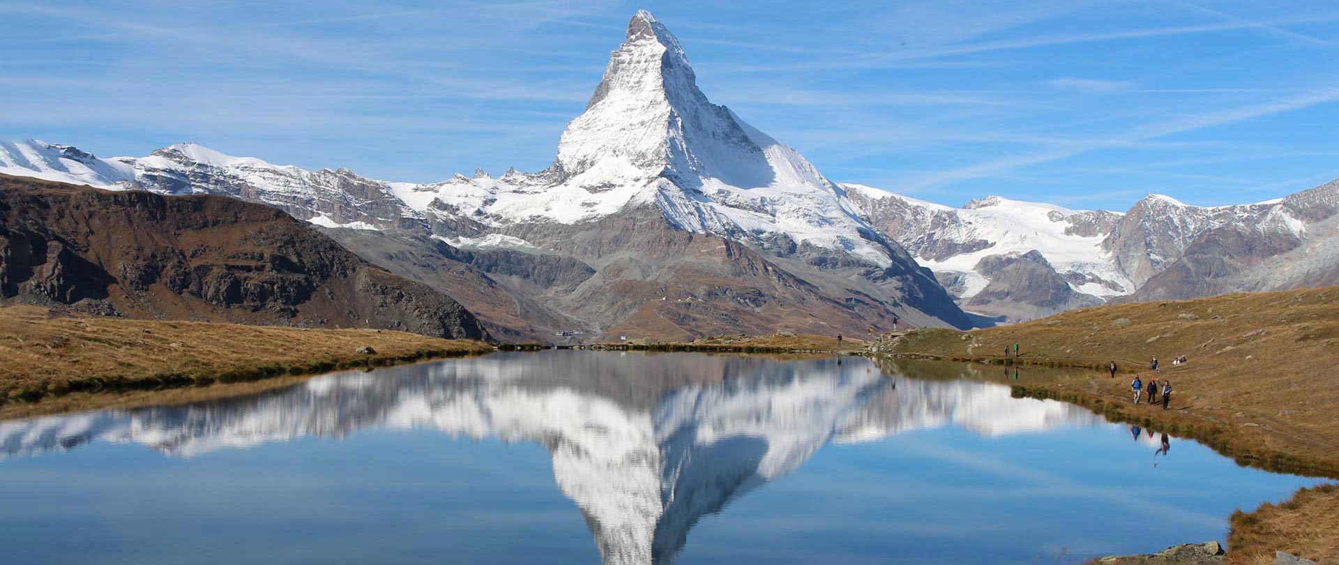 Snow-capped Matterhorn reflected in a calm lake under a clear blue sky, with grassy banks and people along the shoreline—an idyllic Switzerland scene curated by a luxury concierge for an unforgettable experience.