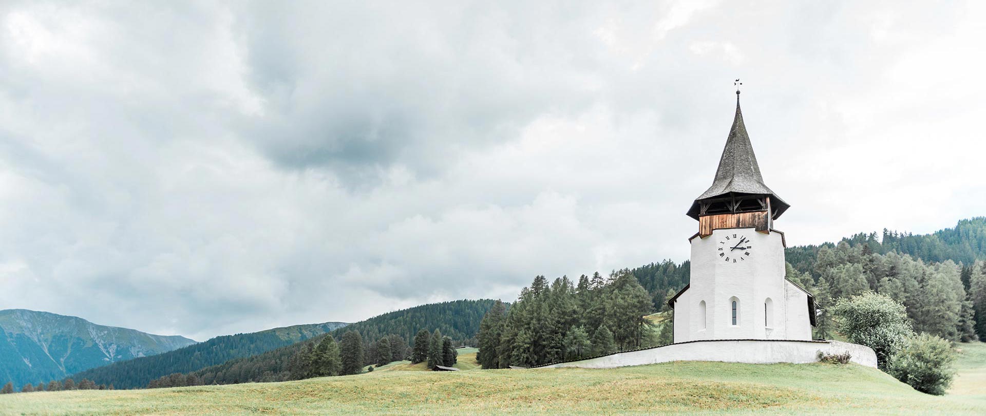 A white church with a clock tower stands on a grassy hill, surrounded by trees and mountains under a cloudy sky—a serene scene reminiscent of the landscapes enjoyed by Swiss luxury services.