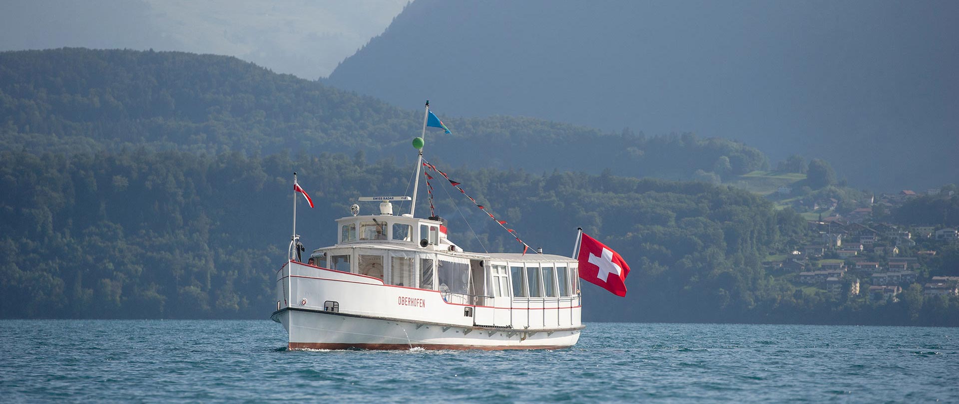 A white passenger boat with a Swiss flag sails on a lake in Switzerland, with forested hills and a small village in the background—an idyllic scene perfect for discovering with expert Swiss Concierge services.