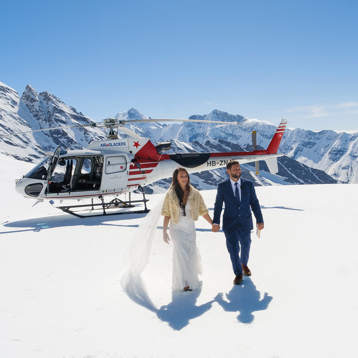 A bride and groom in wedding attire walk on a glacier in the Lauterbrunnen Valley before a helicopter, surrounded by snow-covered mountains—perfect for stunning wedding videography Switzerland has to offer.