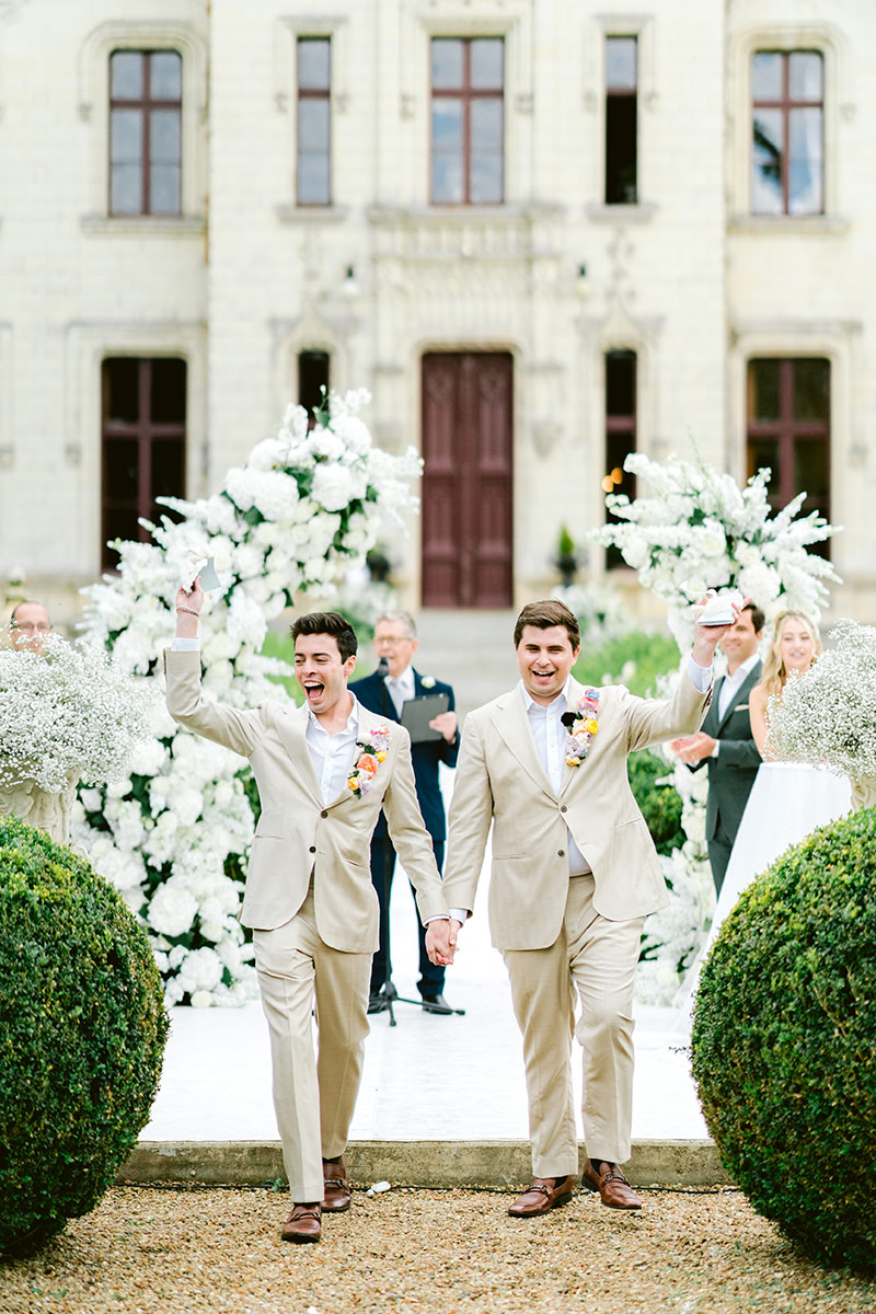Two grooms in beige suits holding hands and celebrating while walking down an outdoor aisle in France, surrounded by floral arrangements and a historic building—the perfect moment for unforgettable wedding videography.