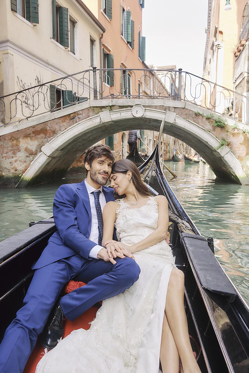 A couple in formal attire sits close together in a gondola on a canal in Venice, with a stone bridge in the background—a perfect scene for destination wedding videography.