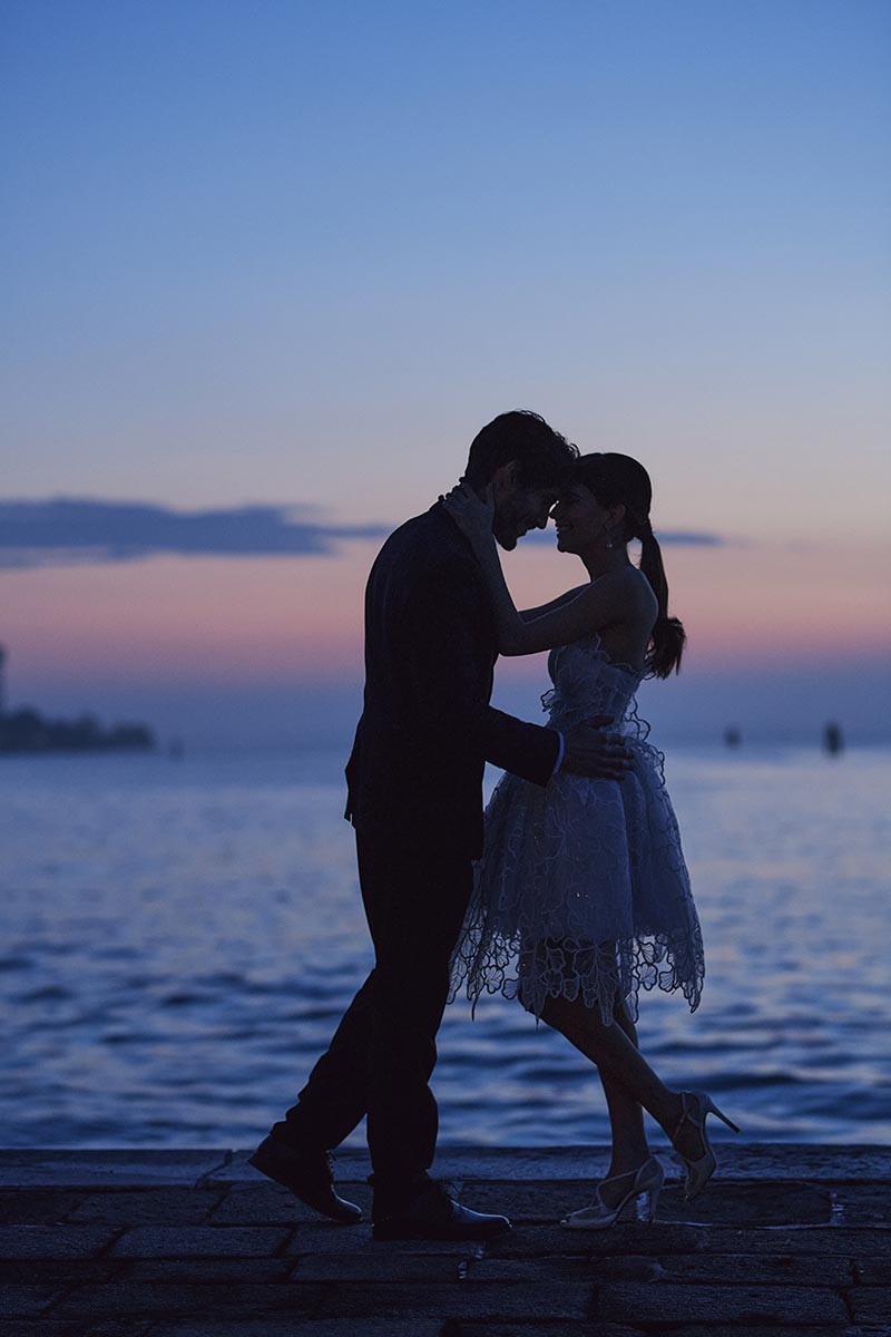 A couple stands close together near a body of water at sunset, silhouetted against a colorful sky—capturing the magic perfect for any wedding video.