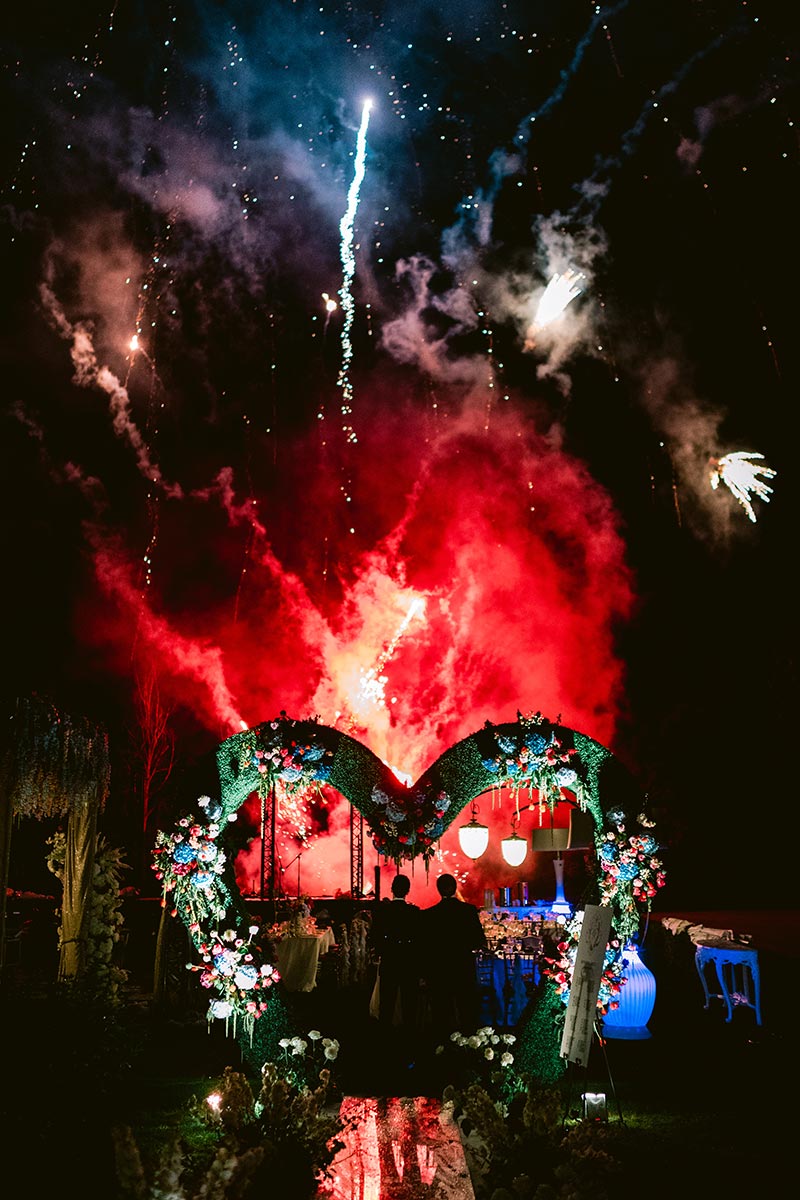 A heart-shaped floral arch and decorated table are illuminated at night as fireworks explode in the sky above, creating colorful smoke and bright light effects—an unforgettable scene for destination videography.
