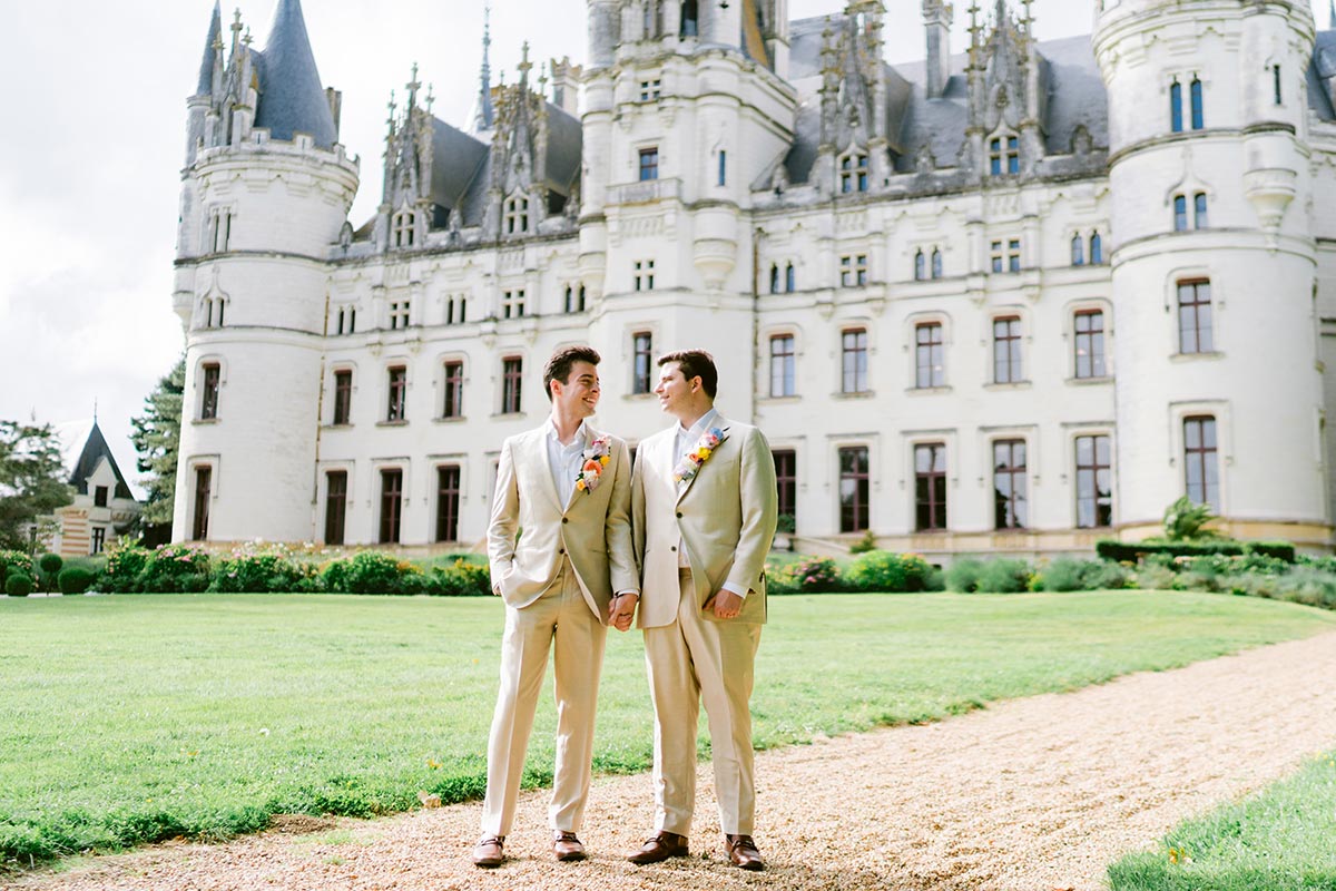 Two men in beige suits holding hands stand on a gravel path before a grand, ornate castle—an idyllic setting for wedding destinations, surrounded by verdant lawns and gardens.