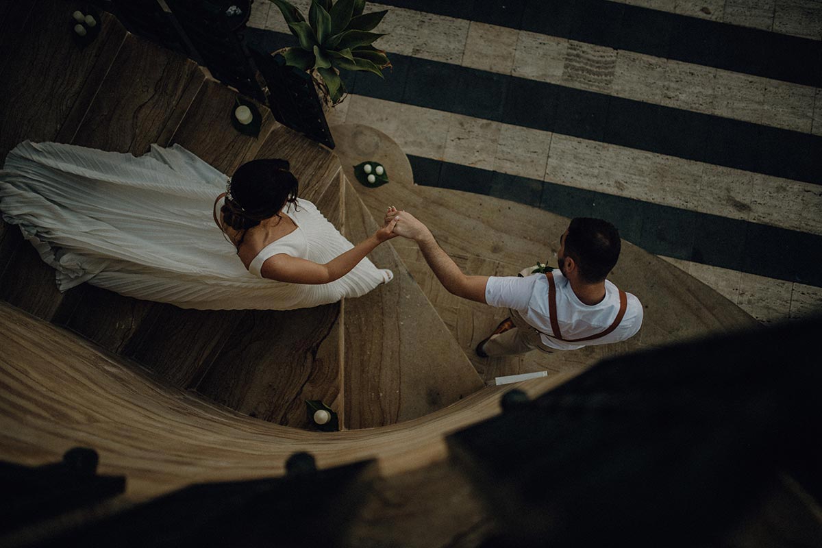 A bride in a white dress holds hands with a man as they walk down a curved wooden staircase, viewed from above..