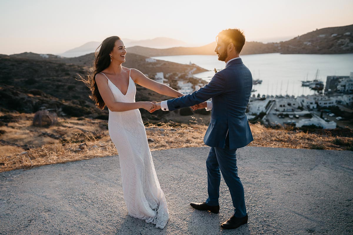 A bride and groom holding hands and smiling at each other outdoors at sunset, with a scenic coastal landscape of IOS in the background