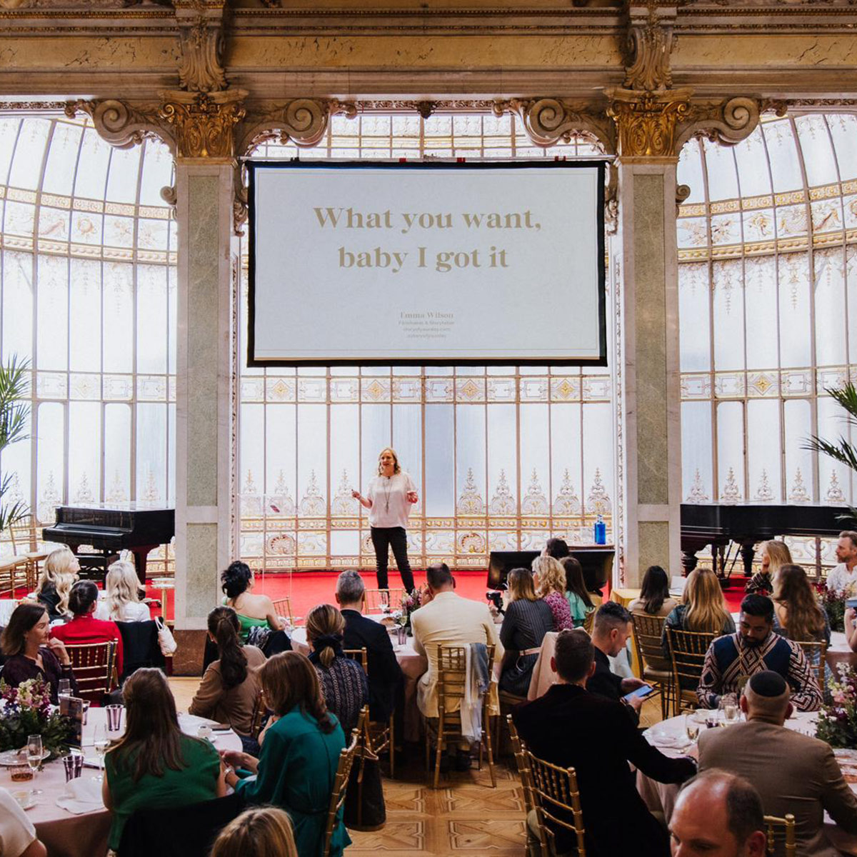 Emma Wilson from Story of Your Day stands on stage in front of a seated audience in an ornate hall, with a large screen displaying the text, "What you want, baby I got it.