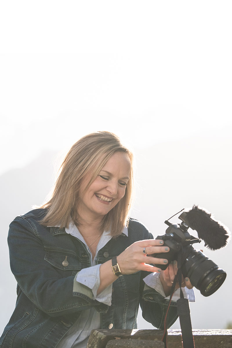 Wedding videographer Emma Wilson smiles in a denim jacket as she adjusts settings on a camera with a microphone attached, outdoors in bright daylight.