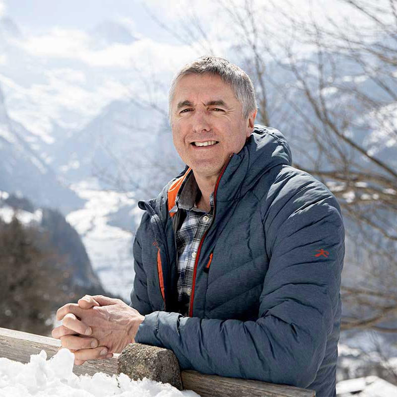 James Wilson est dehors dans un paysage montagneux enneigé, souriant, les mains posées sur une balustrade en bois.