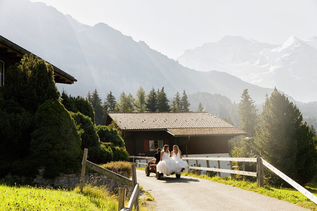 Two brides dressed in white formal attire ride on a small cart up a paved path in a Swiss mountain village, perfect for capturing unforgettable moments with expert wedding videography. Trees and chalet-style buildings line the scenic route.