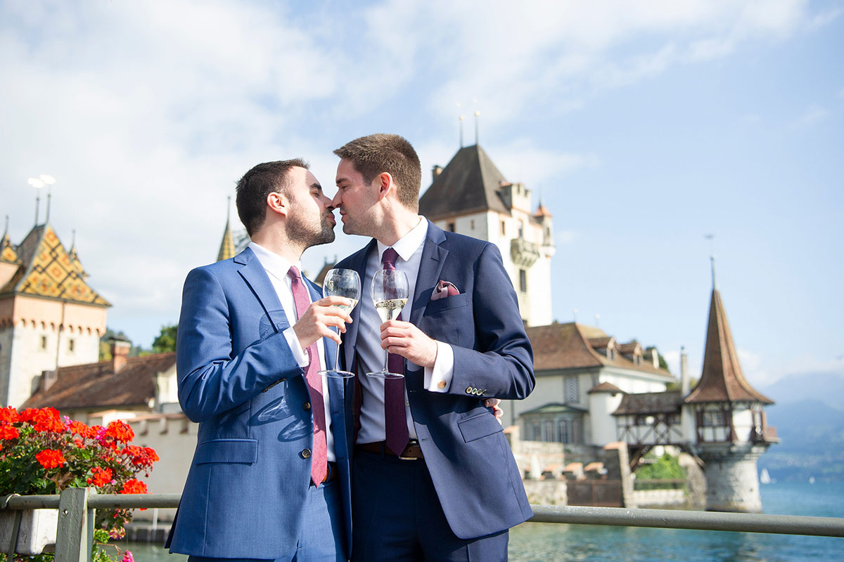Two grooms in front of Schloss Oberhofen clinking wine glasses during their wedding celebration.
