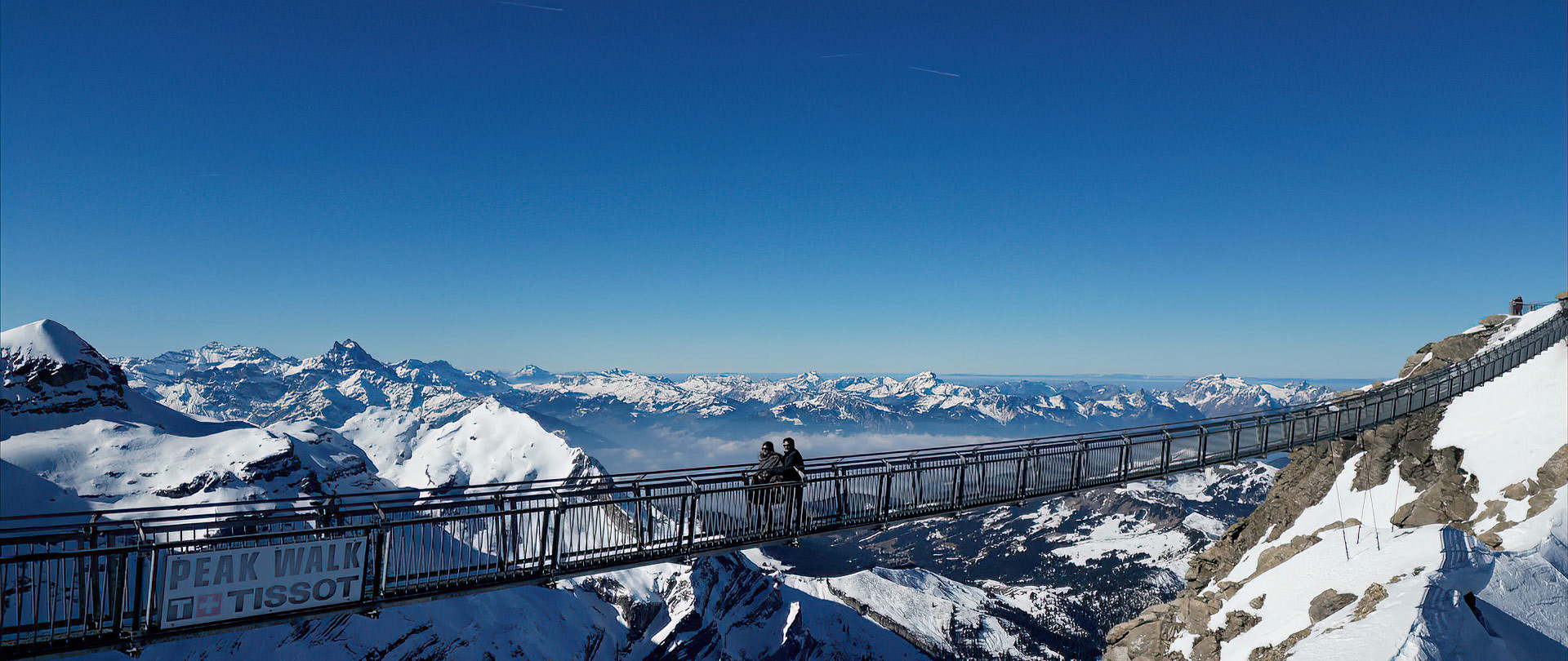 People walk across a suspension bridge high above snowy mountains in Switzerland, with clear blue skies and distant peaks—a breathtaking scene perfect for wedding videography.