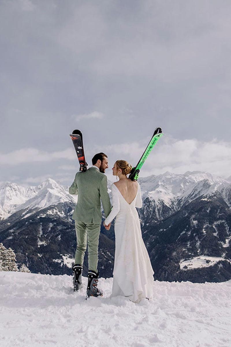 A man in a green suit and a woman in a white dress stand on snow, holding skis and hands, facing majestic snow-covered mountains in Austria—perfect inspiration for your wedding videography.