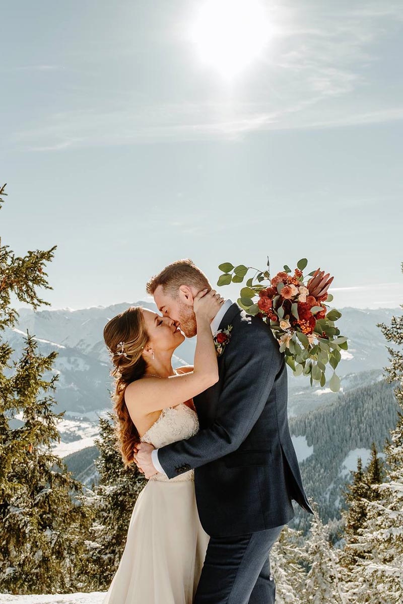 A bride and groom embrace and kiss outdoors in a snowy mountain landscape under a bright sun, with the groom holding a bouquet of flowers—capturing the magic of Austria in this stunning wedding videography moment.